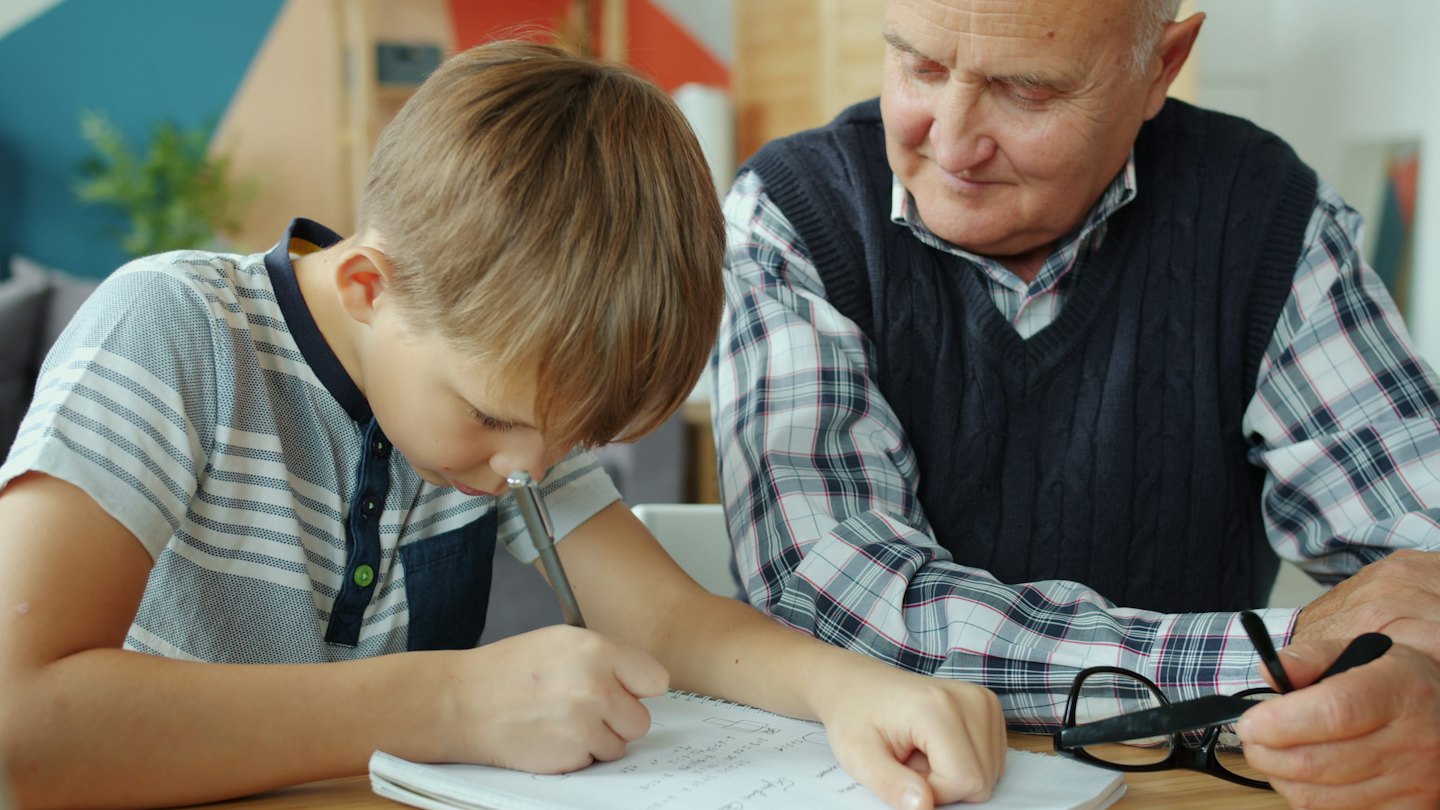 Boy writes in notebook with man watching him.