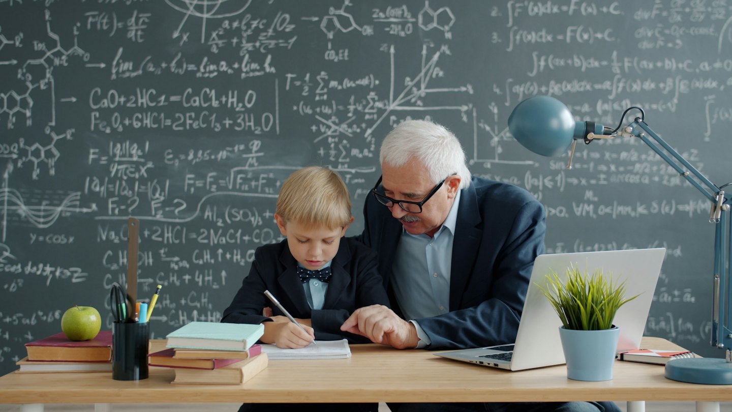 Man helps boy write at a desk with books and a laptop.